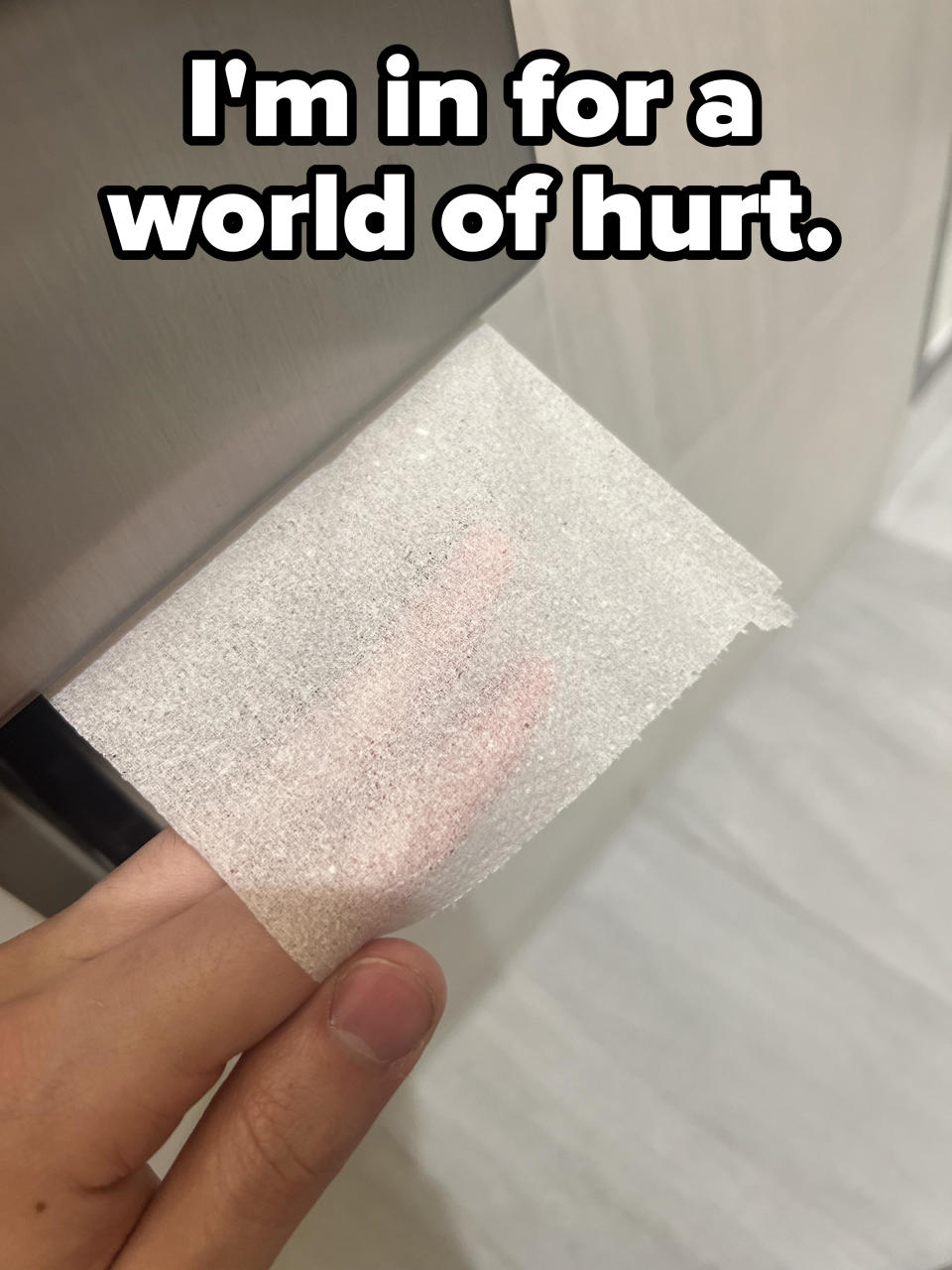 Hand holding a square of toilet paper with a visible pink stain against a restroom wall