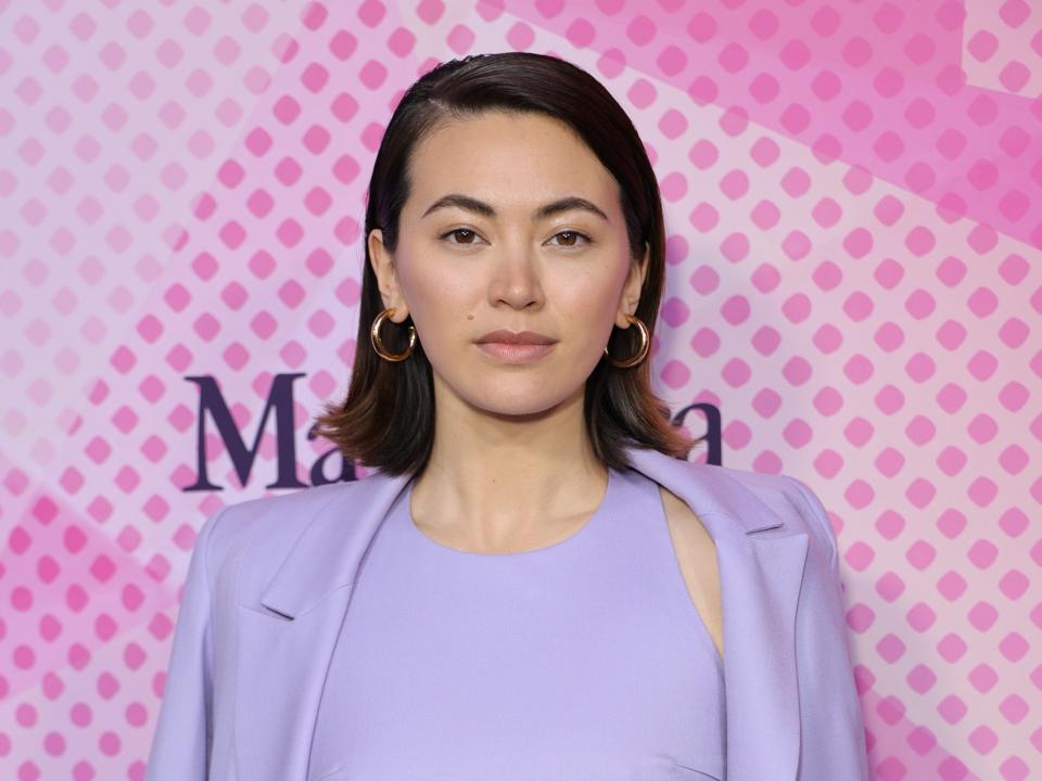 Jessica Henwick in lavender pants, a crop top, and a blazer in front of a pink background