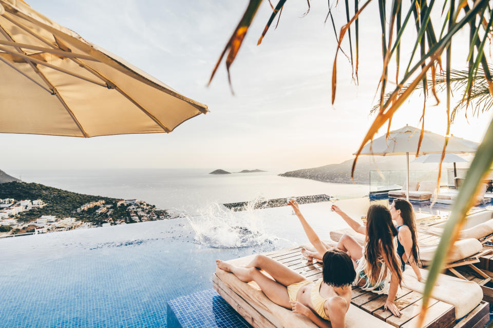 Three women relax on loungers by the edge of an infinity pool overlooking a coastal landscape with islands in the distance; one woman splashes water with her foot