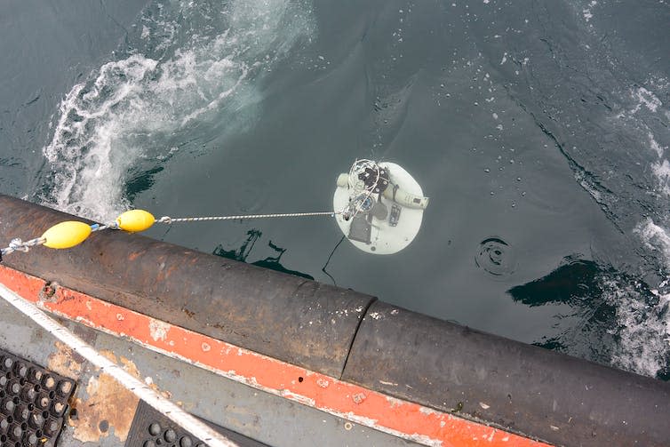 A drum covered in electronic devices is lowered over the side of a boat into the ocean.