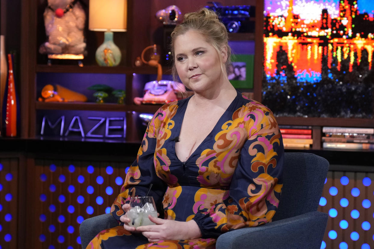 Amy Schumer, wearing a patterned dress with a plunging neckline, sitting while holading glass