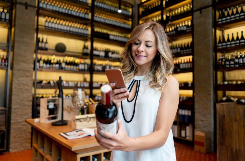 A woman in a wine store, smiling and holding a bottle of wine in one hand while using her smartphone with the other. Shelves filled with wine bottles are visible in the background