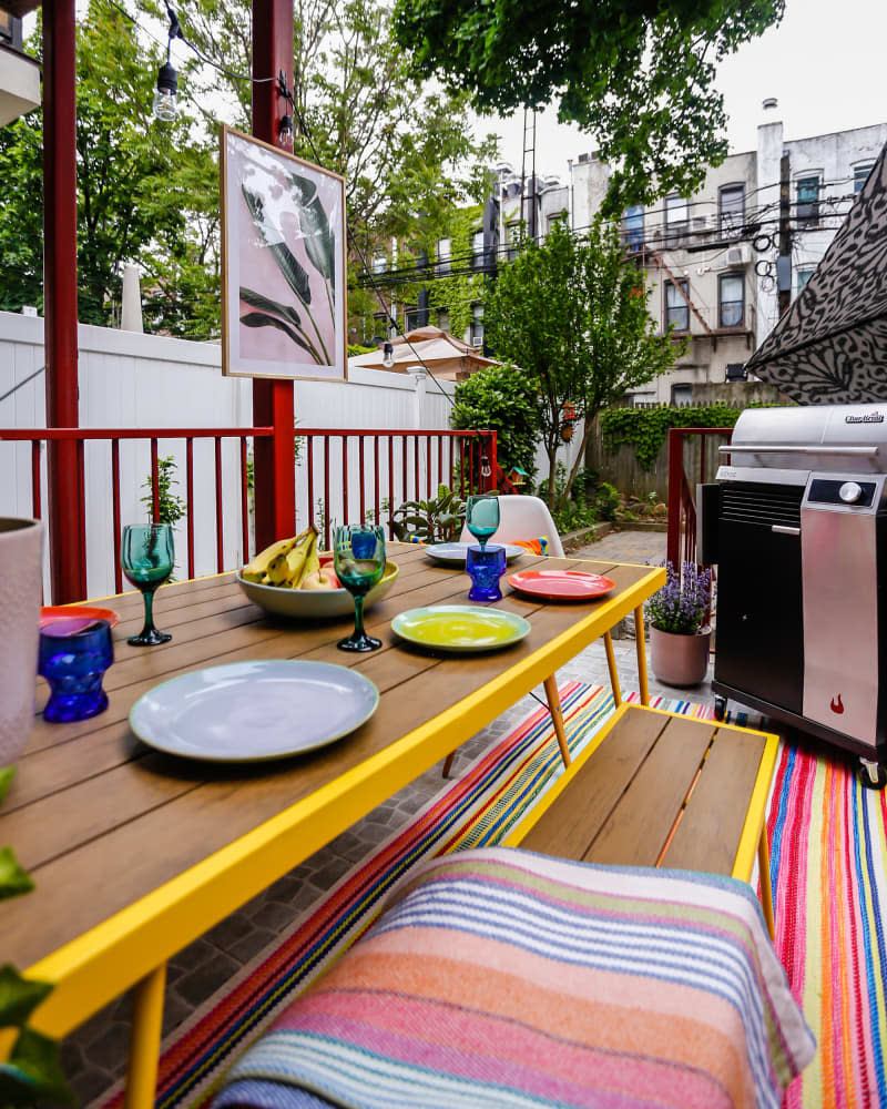 Patio with wood table and bench with yellow trim, colorful rug and dishes on table, and a gas grill