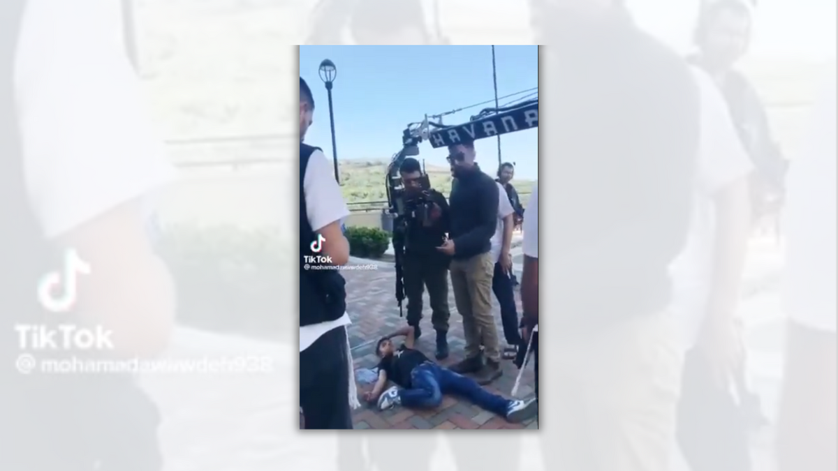 A Palestinian boy lies on the ground. People stand around him . You can see a film camera above him. 