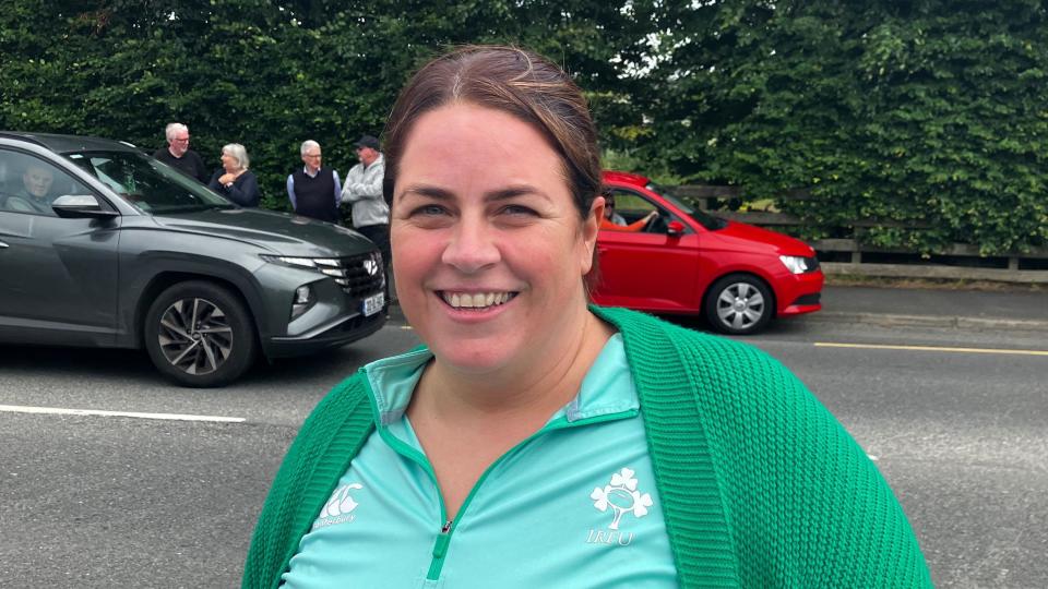 Kara Saunders smiling. She is wearing a green cardigan over a green Irish rugby tracksuit tp. She has brown hair pulled back into a ponytail and is standing in front of a road, on which there are two cars - one red and one dark grey. 