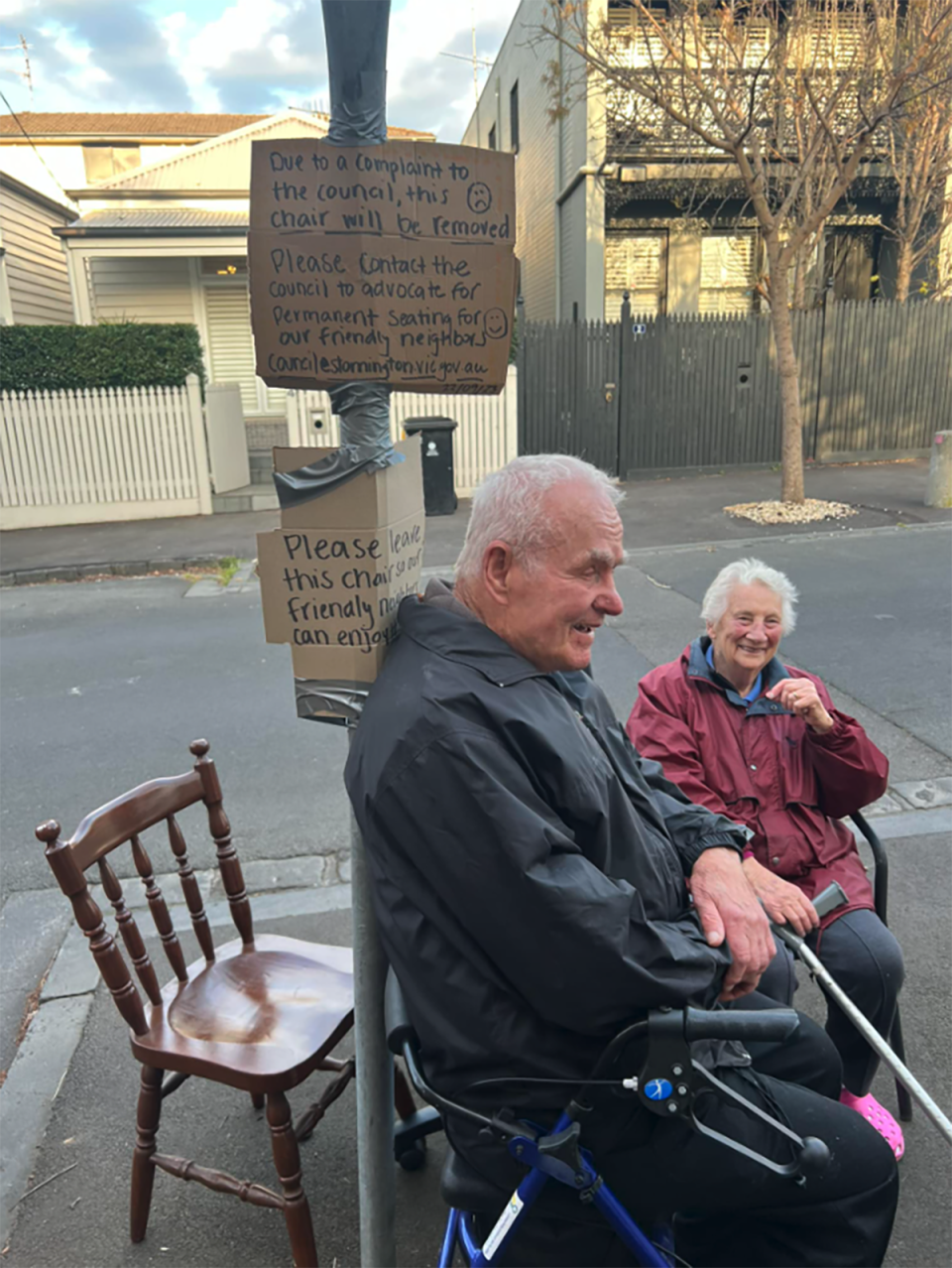 Cuplu de bătrâni așezat pe scaune oferite de localnici pe strada Westbourne din Melbourne.  Se văd semne de carton lipite de un stâlp în spatele ei.