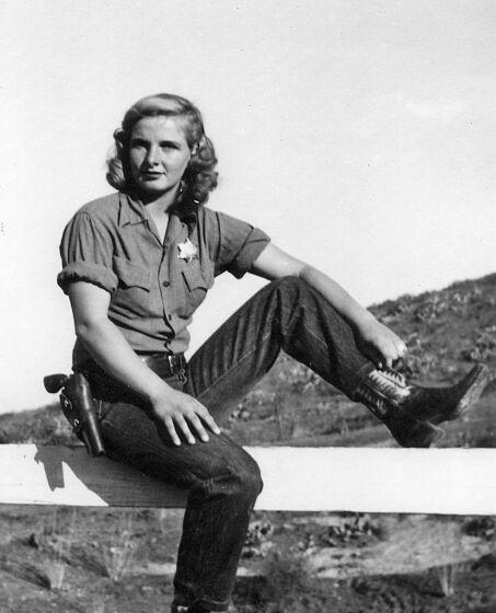 A black-and-white frame of a woman wearing cowboy boots, jeans, belt with a pistol and a sheriff's star, sitting on a beam.