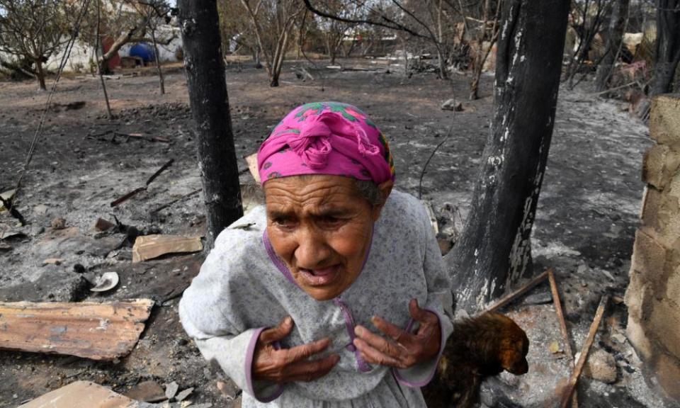 An elderly woman wearing a pink headscarf stands in front of the burned ruins of a house with her hands on her chest