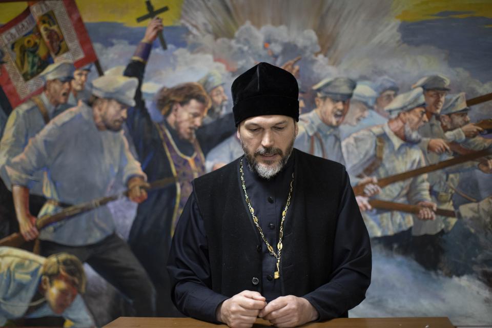 In this photo taken on Tuesday, June 2, 2020, Father Vasily Gelevan speaks during an interview with The Associated Press at the Church of the Annunciation of the Holy Virgin in Sokolniki in Moscow, Russia. The fresco in the background depicts a Russian Orthodox priest walking with a cross in hand alongside Russian army soldiers in a 1904 battle with Japanese troops.In addition to his regular duties as a Russian Orthodox priest, Father Vasily visits people infected with COVID-19 at their homes and hospitals. (AP Photo/Alexander Zemlianichenko)