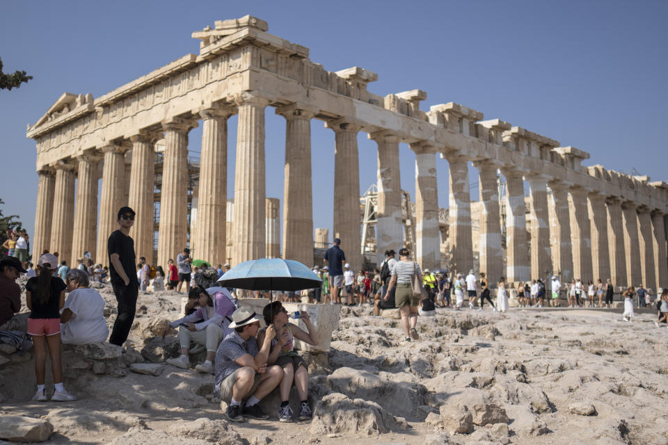 A tourist drinks water as she and a man sit under an umbrella in front of the five century BC Parthenon temple at the Acropolis hill during a heat wave, on Thursday, July 13, 2023. The government has announced emergency measures this week, allowing workers to stay home during peak temperature hours as a heat wave is due to affects most of Greece. (AP Photo/Petros Giannakouris)