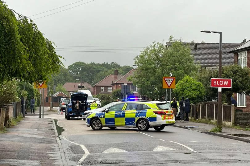 Police car blocking Abbey Road in Beeston, with speed bumps visible in the foreground, red slow signs on lamppost on right, give way signs visible at junction with Marlborough Road and other people on pavement on right, left and sitting in boot of blue car behind pollice car