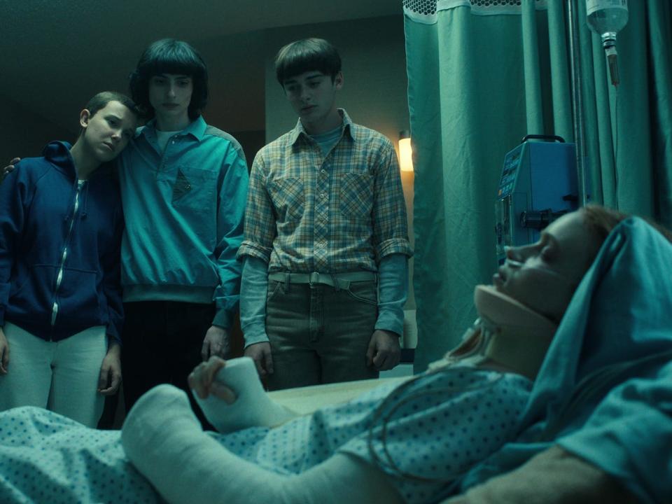 Four teenagers (Lucas, Eleven, Mike, and Will) stand over the unconscious body of their friend, Max, in a hospital room.