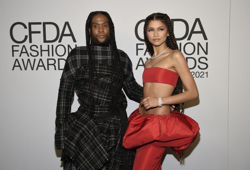 A man with braids in a dark plaid gown and Zendaya in a bright red bandeau and puffy skirt