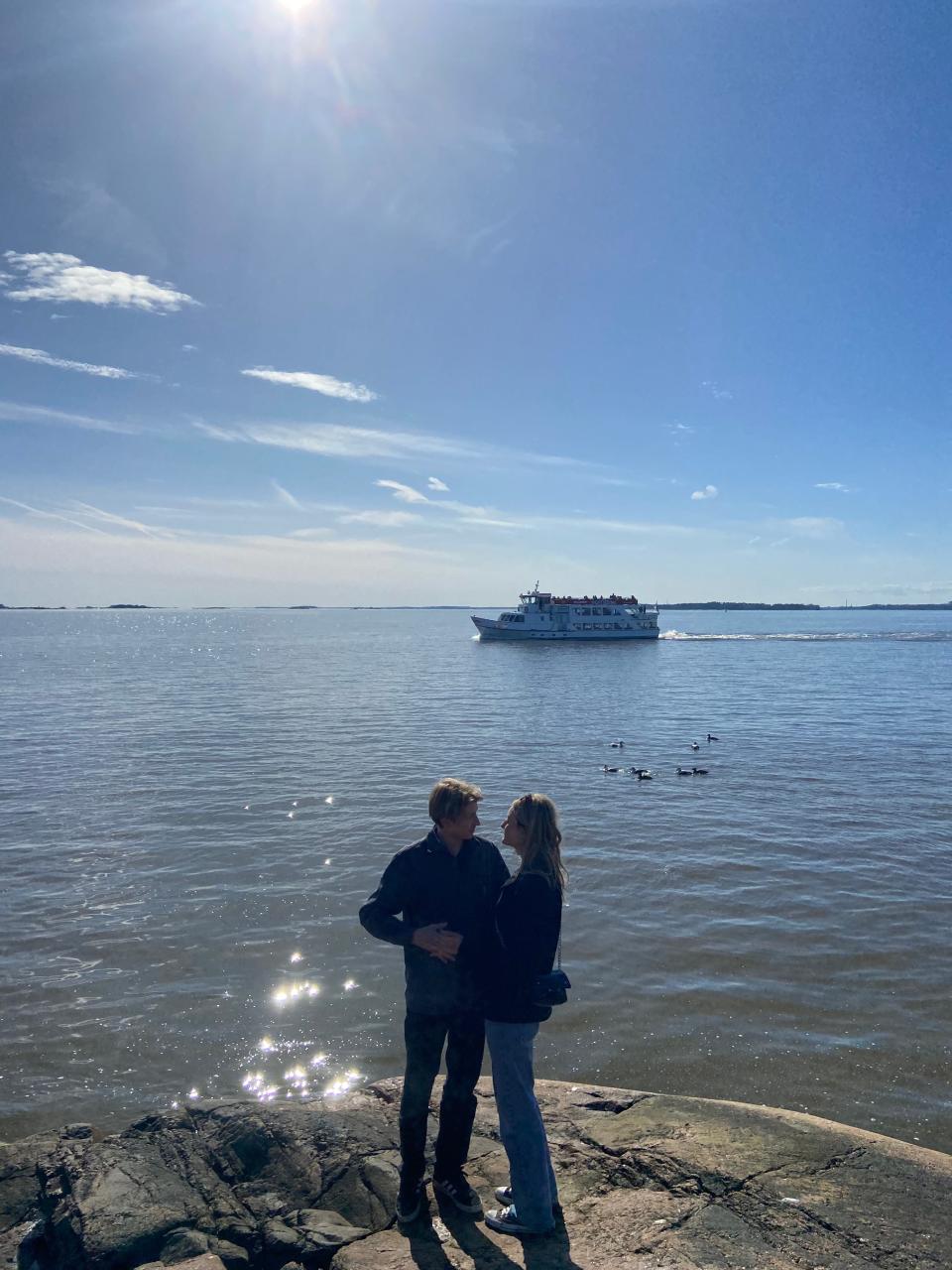 Oliver Russell and Helena Tomaszewska. embrace on a rocky outcrop in front of a large body of water where a boat sails past.