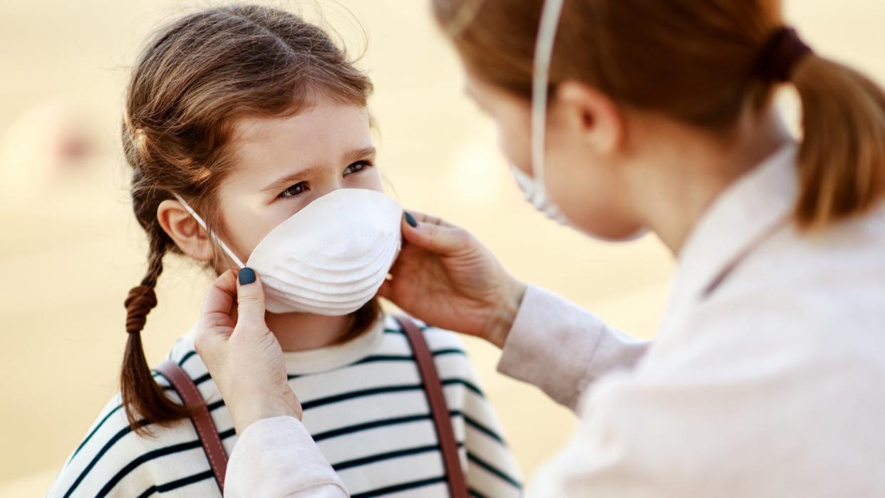 From above adult woman putting on medical mask of little girl while standing on city street during coronavirus pandemic.