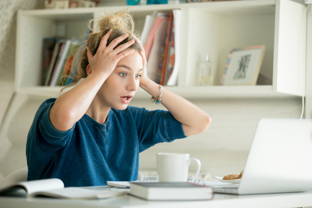 Portrait of an attractive woman at the table with cup and laptop, book, notebook on it, grabbing her head. Bookshelf at the background, concept photo