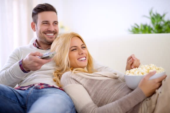 A smiling young couple, leaning on each other and a couch with popcorn and a TV remote close at hand.