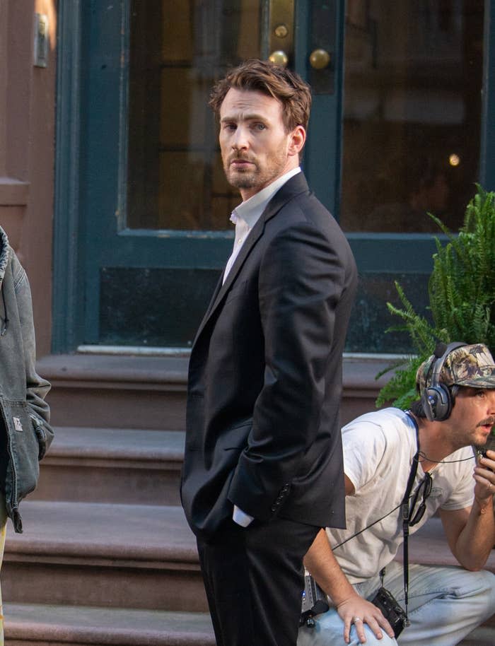 Chris Evans stands on a street in a suit, filming a scene, with a crew member in a hat and headset crouching nearby