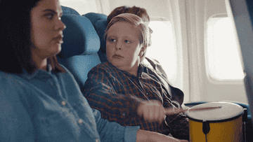 Kid tapping on a drum on a plane next to a woman who appears visibly annoyed