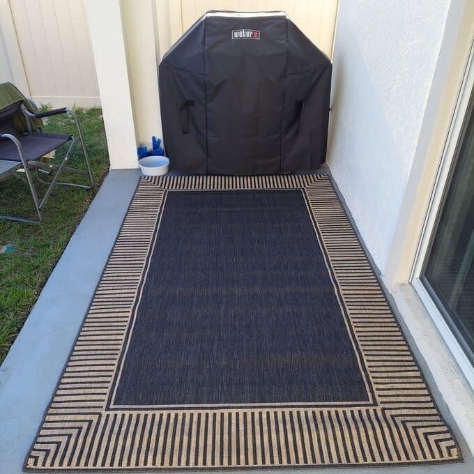 A Weber grill under a cover next to a chair on a patio with a striped outdoor rug