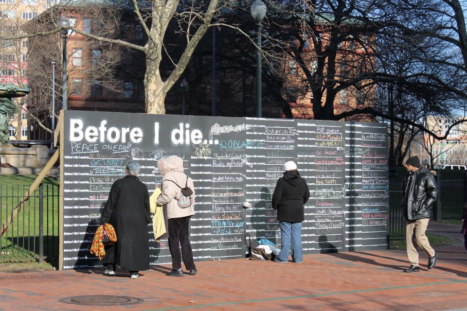 In this Nov. 29, 2013 photo, people finish the sentence “Before I die . . .” on a large blackboard in Providence, R.I. Since 2011, 400 such walls have gone up in the United States as well as in 60 other countries. The global phenomenon has oversize blackboards, painted on buildings and freestanding displays that invite those passing by to complete the sentence. (AP Photo/Michelle Smith)