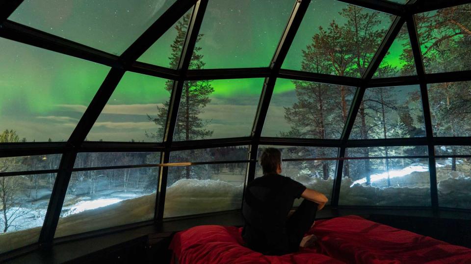 A man sits on a red bed in a glass guest tent in Finland, observing the green sky (Northern Lights) and stars