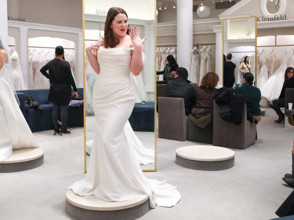 A woman holds up her ring finger while she wears a wedding dress in a bridal boutique.