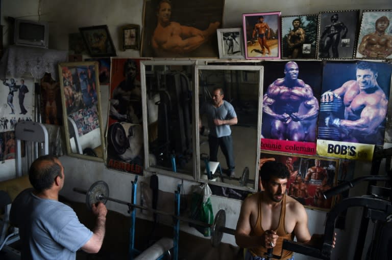 Men grunt and sweat their way through a workout beneath the watchful eye of a young Arnold Schwarzenegger, whose muscle-bound image hangs from the wall