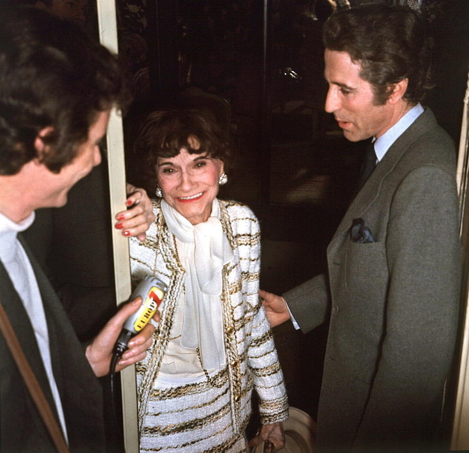 I don't know who is in this image. An elderly woman in a striped jacket and white dress is smiling while holding the door, speaking to two men. One man is holding a microphone