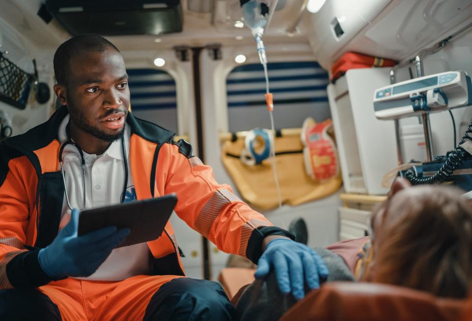 A paramedic in an ambulance holds a tablet while assisting a patient lying down, providing urgent medical care