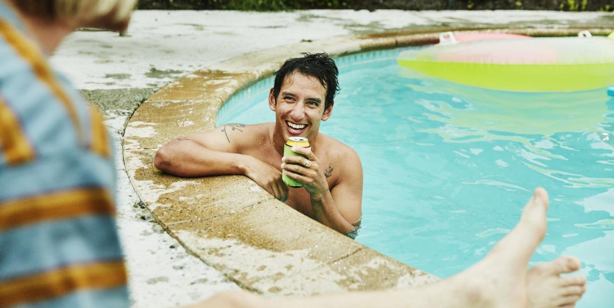 laughing man with drink in backyard pool in discussion with friend on summer evening