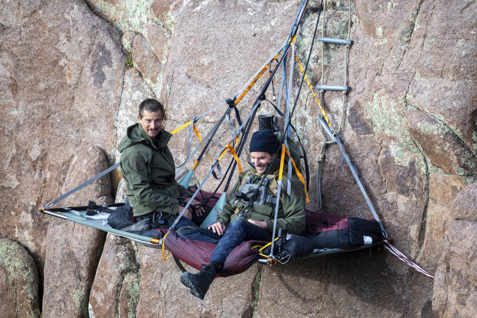 CORRECTS POSITION OF COOPER TO RIGHT - This image released by Nat Geo shows Bradley Cooper, right, and Bear Grylls on a paraledge hung off the edge of Pathfinder Canyon in Wyoming, in a scene from “Running Wild with Bear Grylls: The Challenge," premiering on July 9. (Jeff Ellingson/Nat Geo via AP)