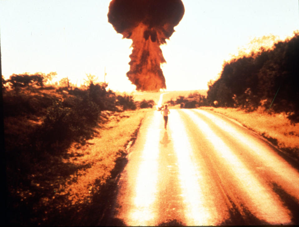 A historical photo showing a large mushroom cloud in the distance with a person viewed from behind witnessing the event