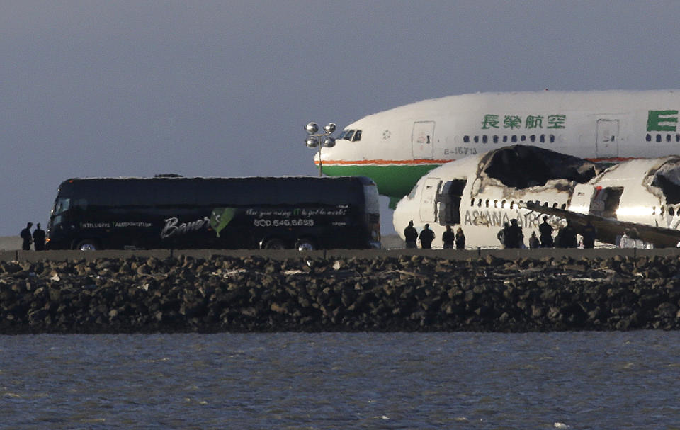 A group of people stand in front of the wreckage of Asiana Flight 214, which crashed on Saturday, July 6, 2013, as one of three buses that were reported to be carrying passengers and family members are parked next to it on a tarmac at San Francisco International Airport in San Francisco, Wednesday, July 10, 2013. Two passengers were killed and many others were injured in the crash. (AP Photo/Jeff Chiu)