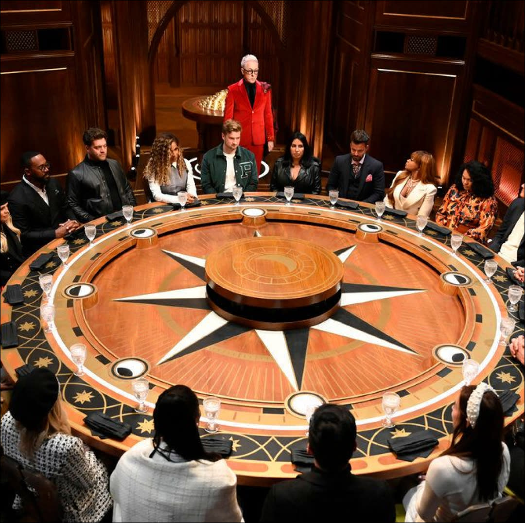  A group of people sit around a round table decorated with a star motif, as alan cumming stands among them wearing a red suit, in 'the traitors'. 