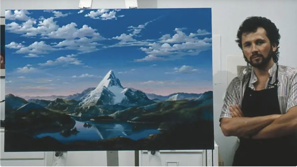 Dario with arms folded next to a painting of snowcapped mountains and clouds in the sky