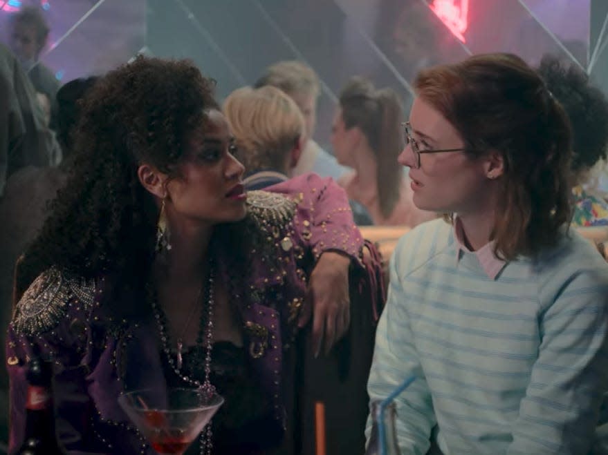 two women look at each other in a club, sitting close together. the one on the left has curly hair and a jacket emblazoned with rhinestones, while the one on the right has on a collared, striped shirt and is wearing her straight hair half back