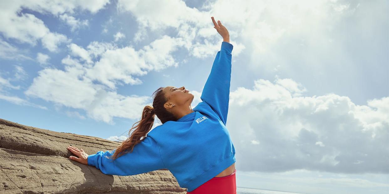 women posing wearing red bikini bottoms, a blue hoodie, one arm stretched to the sky with the other on a rock with a view of the ocean behind her