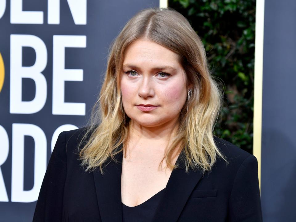 Merritt Wever in front of a blue background and leafy bush wearing a black blazer and black shirt