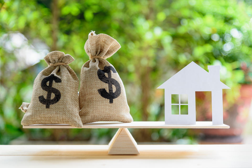 Home loan / reverse mortgage or transforming assets into cash concept : House paper model , US dollar hessian bags on a wood balance scale, depicts a homeowner or a borrower turns properties into cash