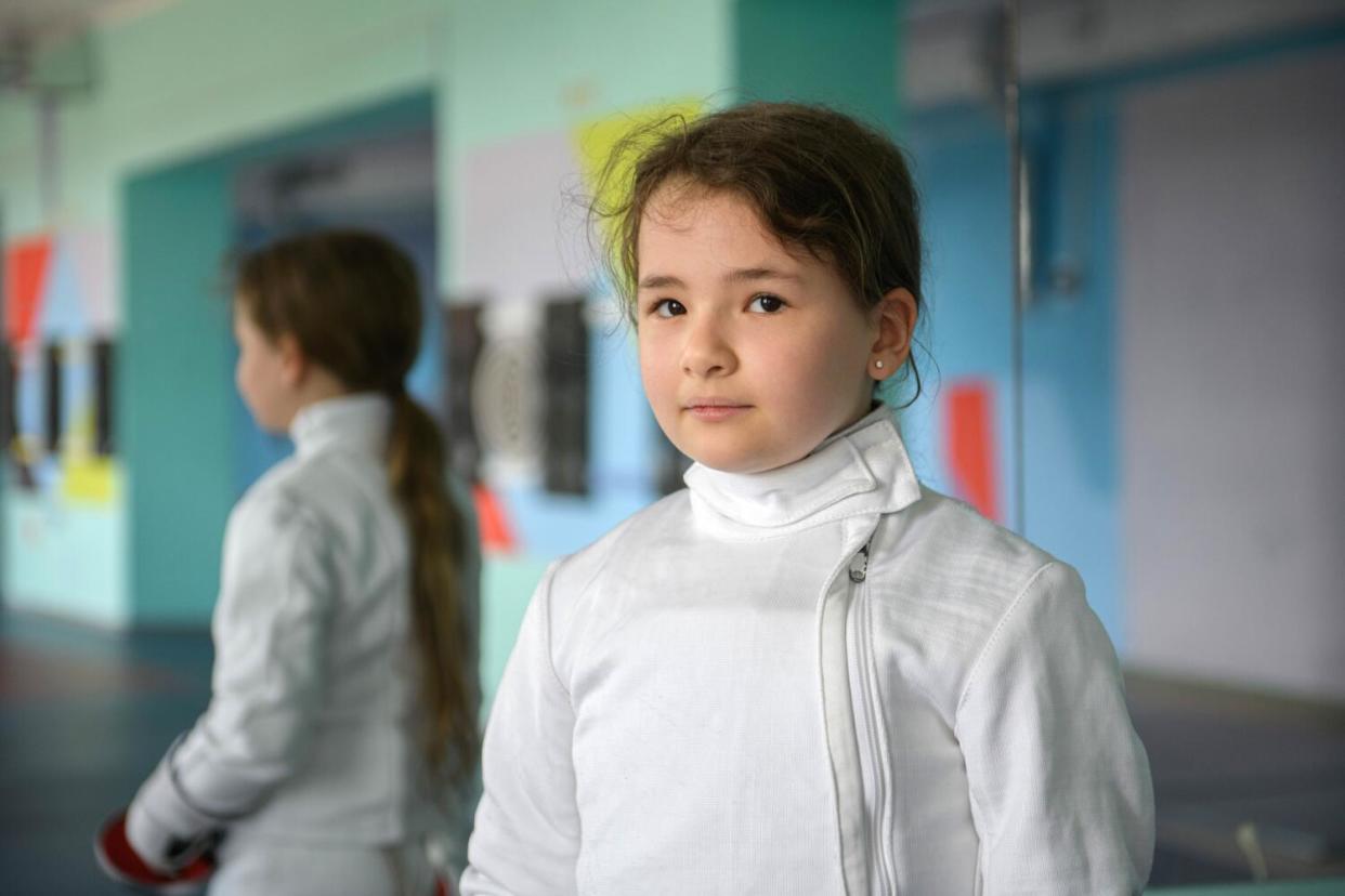 Two girls in white fencing suits stand in a studio.