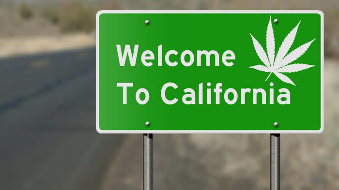 A green highway sign that reads, Welcome to California, with a white cannabis leaf in the upper right-hand corner.
