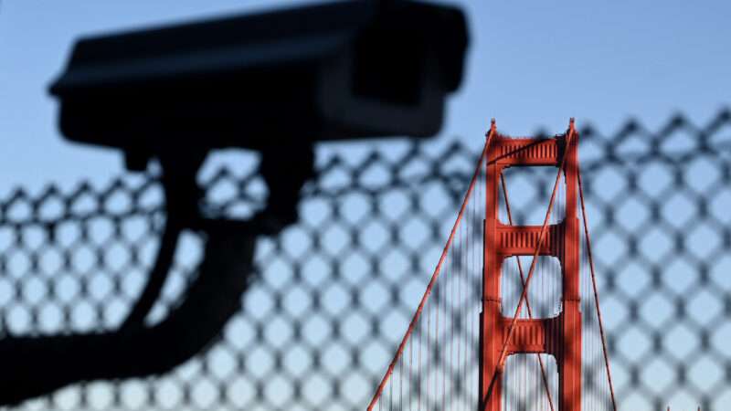 A surveillance camera in the foreground with the Golden Gate Bridge visible in the background, separated by a chain link fence.