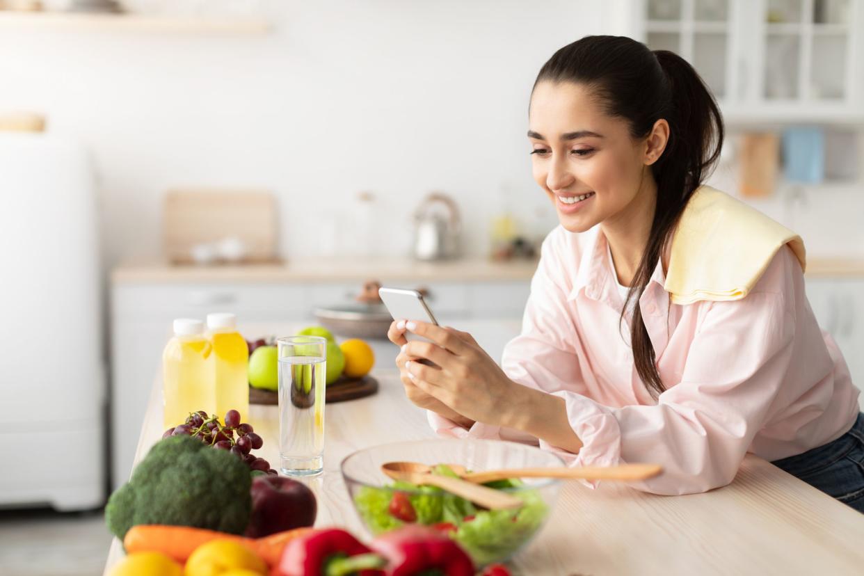 People And Technology Concept. Cheerful smiling millennial woman using mobile phone, standing in kitchen and leaning on counter, cheking social media or searching for new recipe, free copy space