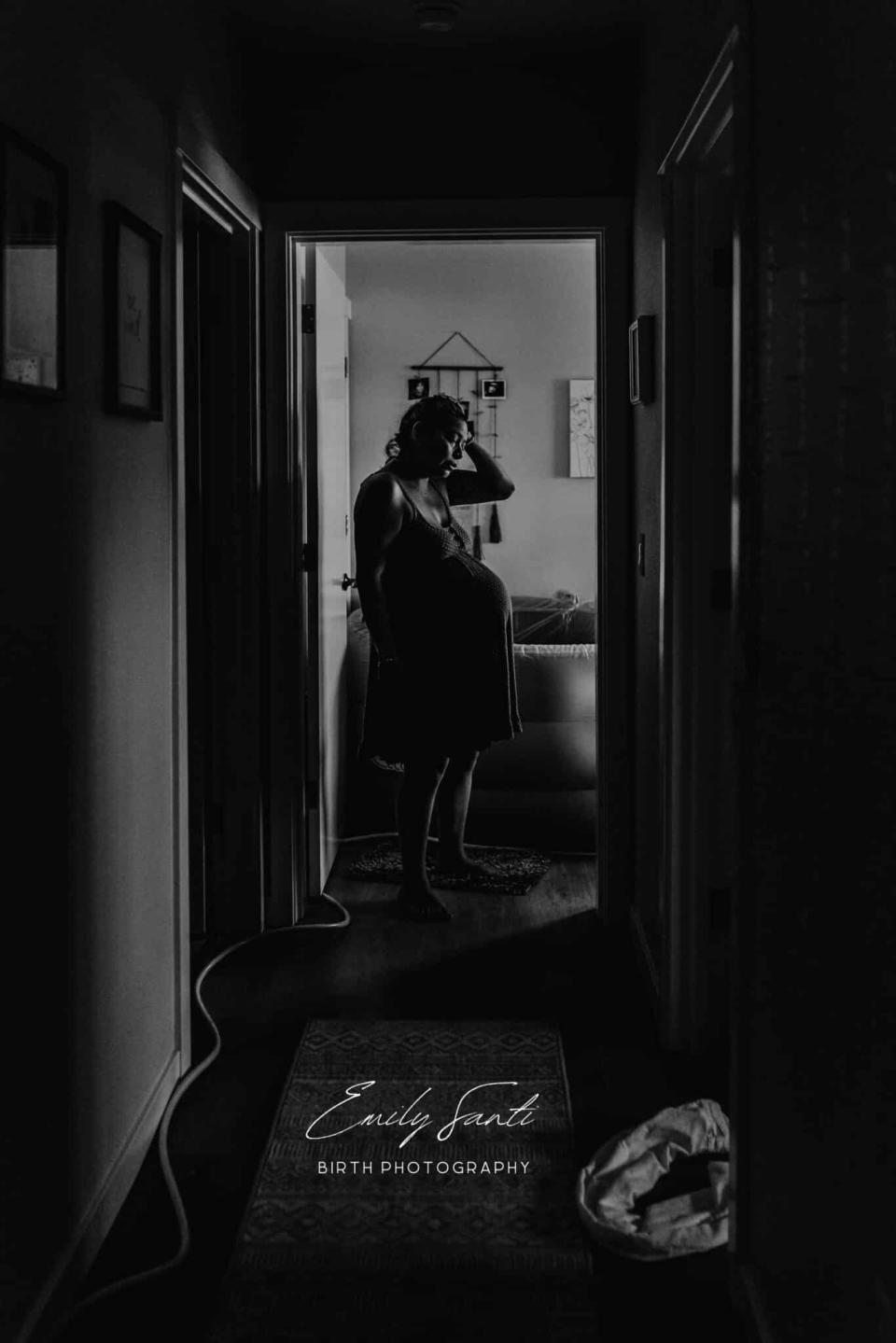 Pregnant person stands in doorway, silhouetted, touching belly; peaceful atmosphere. Photographer's watermark present