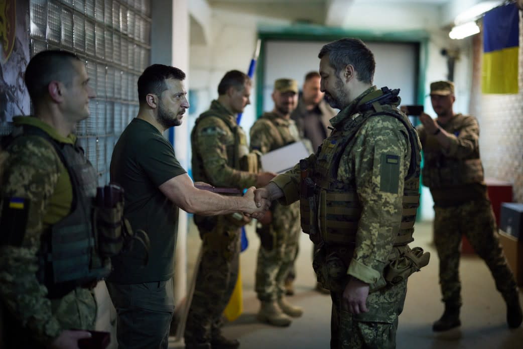 Volodymyr Zelensky, wearing a T-shirt and cargo pants, looks into the eyes of a service member as he shakes the hand, near five other service members, all wearing battle fatigues.