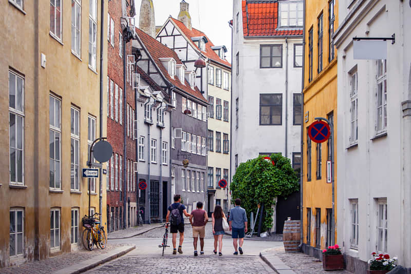 Beautiful bright cityscape in perspective. View on cobbled street with bicycles. Narrow street with colorful buildings in old historic center of Copenhagen. Red tiled roofs. Four people men go forward