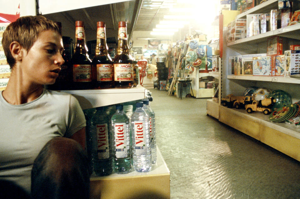 Person sitting by shelves filled with Angostura bitters bottles and Vittel water bottles in a brightly lit store with toys and other items