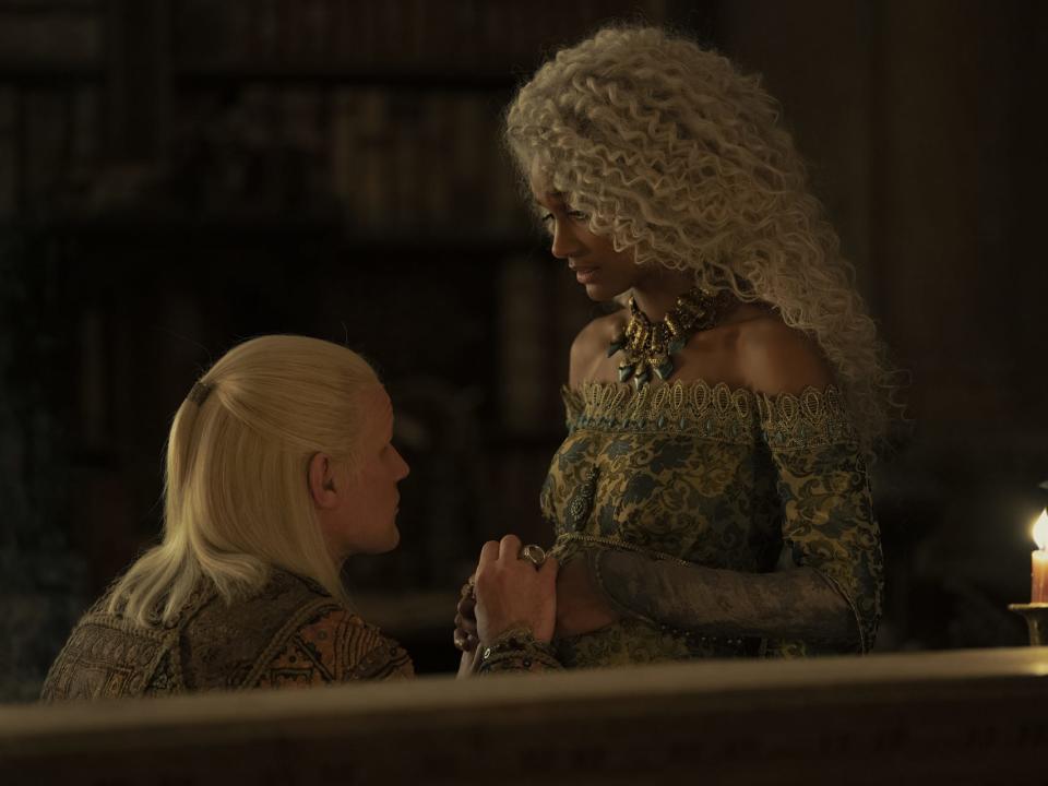 matt smith as daemon targaryen, kneeling in front of his pregnant wife, laena targaryen, played by nanna blondell. daemon has his hands over laena's belly, and she's looking down at him