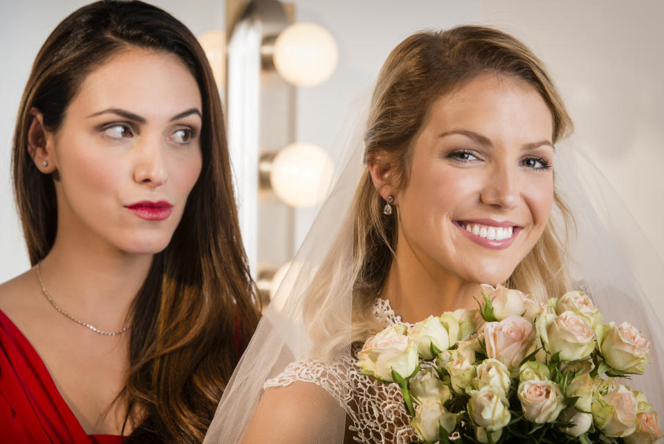 The bride, holding a bouquet of roses, smiles while a bridesmaid in a red dress looks at her with a surprised expression.